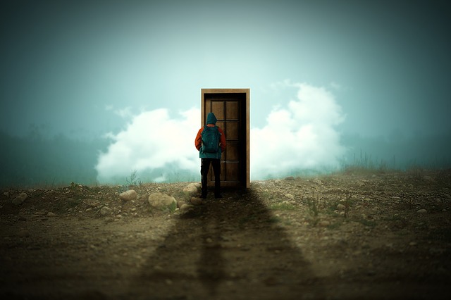 Muž s ruksakom a v bunde stojí pred dverami na lúke pri zamračenom nebi.jpg